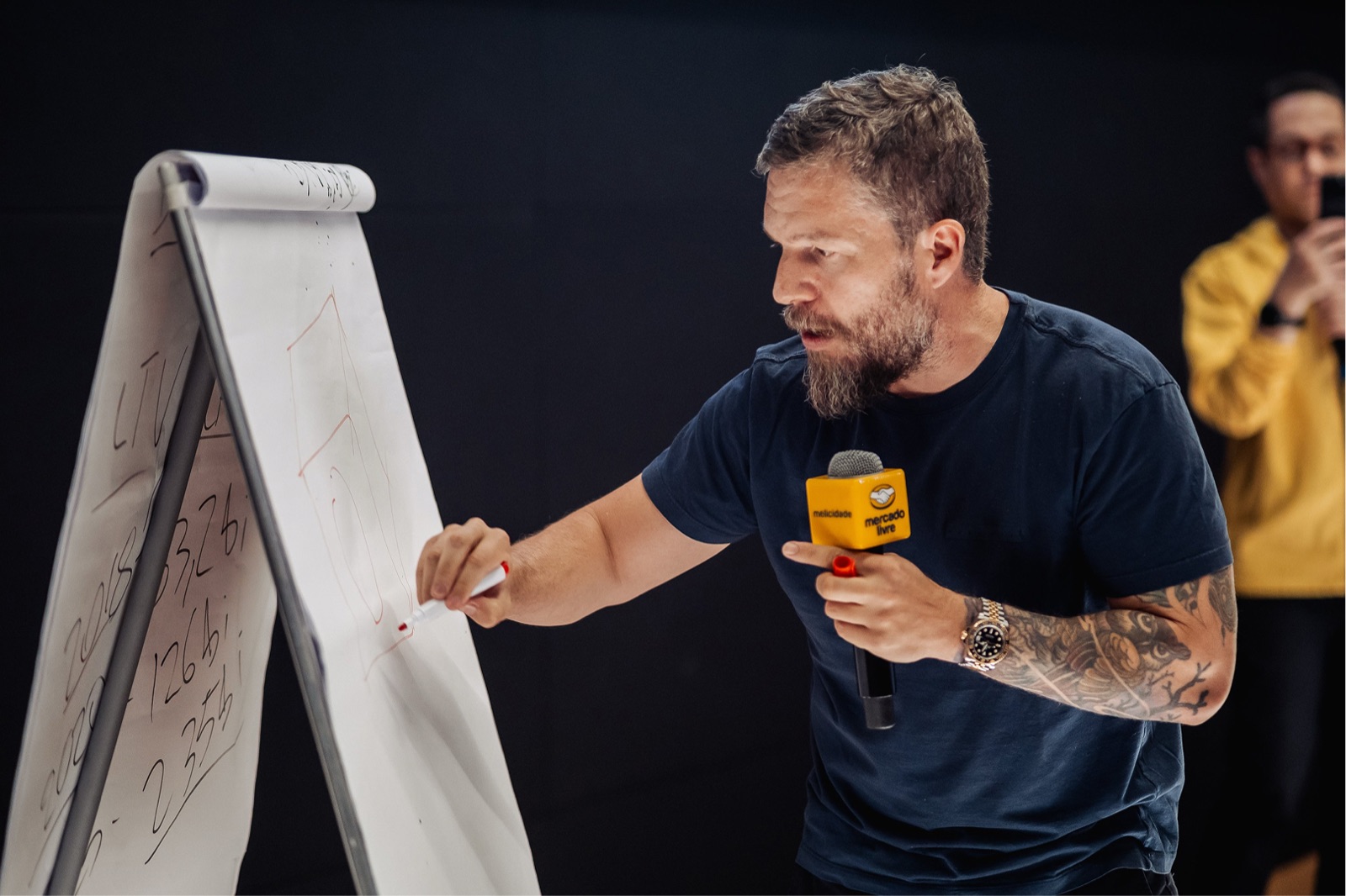 Dhiego Rosa escrevendo números em um flipchart, segurando um microfone do Mercado Livre